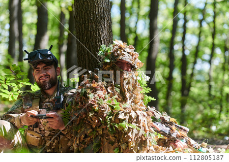 A skilled sniper and a soldier operating a drone with VR goggles strategize and observe the military action while concealed in the forest 112805187