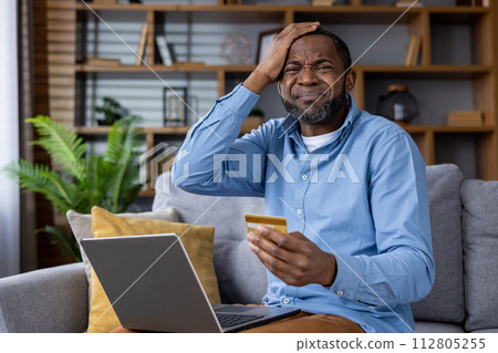 Adult male at home looking at his laptop with a concerned expression, holding a credit card, potentially a victim of online fraud. 112805255