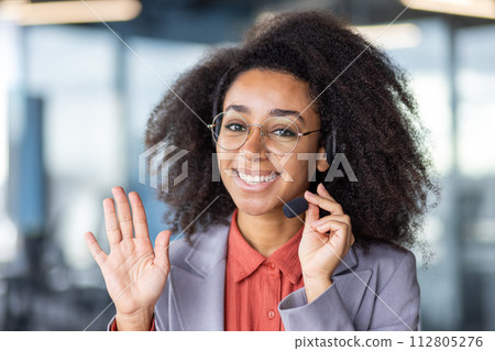 Smiling young woman wearing headset in office setting, waving and providing excellent customer service with a friendly demeanor. Smiling young woman wearing headset in office setting, waving and providing excellent customer service with a friendly demeanor. 112805276