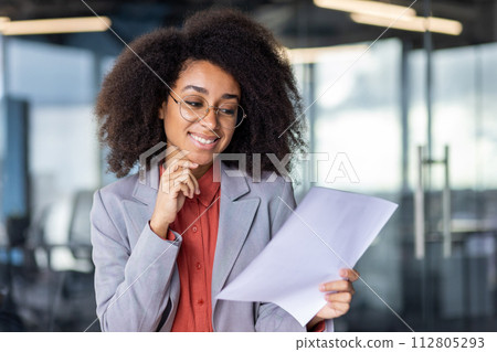A professional young woman with curly hair wearing glasses reviews paperwork with a thoughtful expression in a modern office setting. 112805293