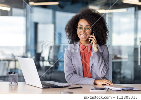 A young professional woman engages in a phone conversation at her office desk surrounded by technology and modern office decor. 112805313