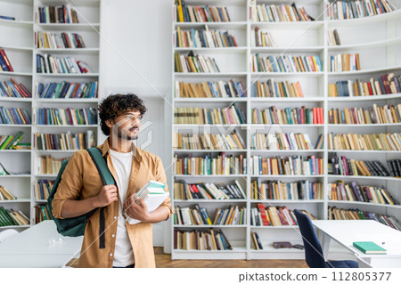 Confident male student holding textbooks in a contemporary library setting, looking away thoughtfully amidst bookshelves. Confident male student holding textbooks in a contemporary library setting, looking away thoughtfully amidst bookshelves. 112805377