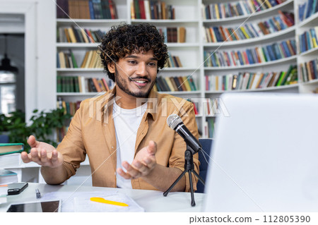 An engaging podcaster in a casual setting, discussing topics live with a microphone and laptop in front of bookshelves. 112805390