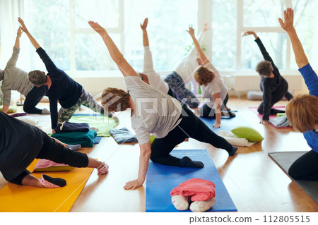 A group of senior women engage in various yoga exercises, including neck, back, and leg stretches, under the guidance of a trainer in a sunlit space, promoting well-being and harmony 112805515