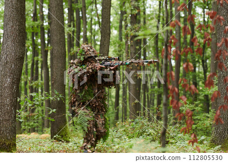 A highly skilled elite sniper, camouflaged in the dense forest, stealthily maneuvers through dangerous woodland terrain on a covert and precise mission 112805530