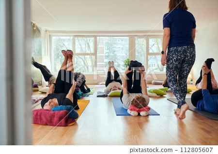 A group of senior women engage in various yoga exercises, including neck, back, and leg stretches, under the guidance of a trainer in a sunlit space, promoting well-being and harmony 112805607