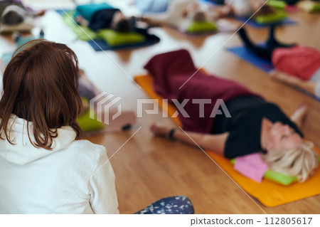 A group of senior women engage in various yoga exercises, including neck, back, and leg stretches, under the guidance of a trainer in a sunlit space, promoting well-being and harmony 112805617