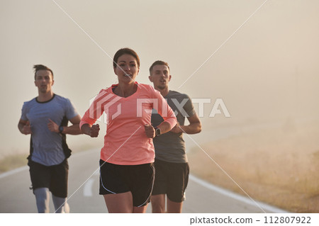 A group of friends, athletes, and joggers embrace the early morning hours as they run through the misty dawn, energized by the rising sun and surrounded by the tranquil beauty of nature A group of friends, athletes, and joggers embrace the early morning hours as they run through the misty dawn, energized by the rising sun and surrounded by the tranquil beauty of nature 112807922