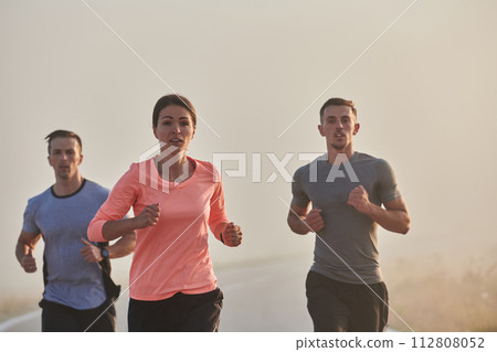 A group of friends, athletes, and joggers embrace the early morning hours as they run through the misty dawn, energized by the rising sun and surrounded by the tranquil beauty of nature A group of friends, athletes, and joggers embrace the early morning hours as they run through the misty dawn, energized by the rising sun and surrounded by the tranquil beauty of nature 112808052