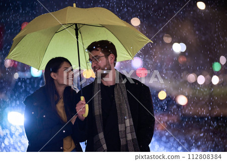 In the romantic ambiance of a rainy night, a happy couple walks through the city, sharing tender moments under a yellow umbrella, surrounded by the glistening glow of urban lights 112808384