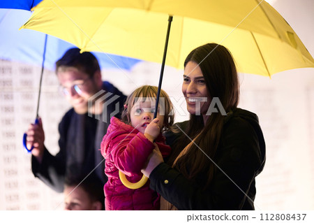 In the midst of a rainy urban night, a happy couple takes their children on a stroll through the city streets, heading towards the cinema for a delightful family movie outing In the midst of a rainy urban night, a happy couple takes their children on a stroll through the city streets, heading towards the cinema for a delightful family movie outing 112808437