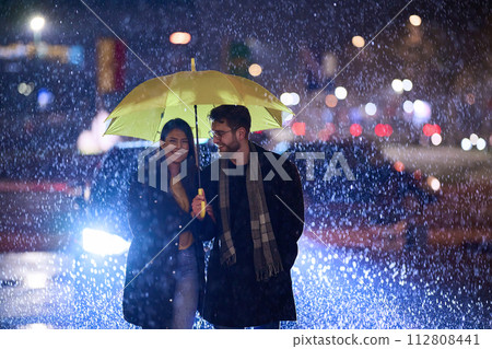 In the romantic ambiance of a rainy night, a happy couple walks through the city, sharing tender moments under a yellow umbrella, surrounded by the glistening glow of urban lights 112808441