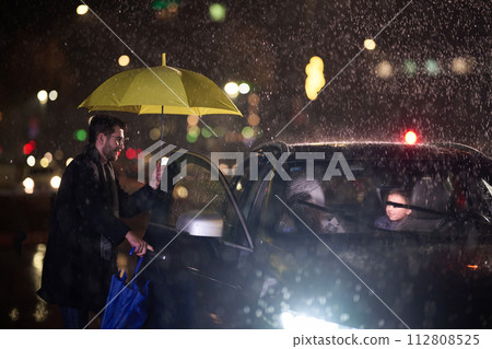A family man lovingly opens the car for his family in rainy weather, sheltering them with an umbrella A family man lovingly opens the car for his family in rainy weather, sheltering them with an umbrella 112808525
