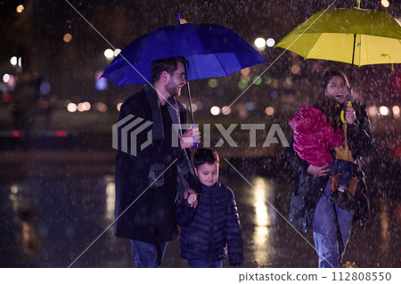 In the midst of a rainy urban night, a happy couple takes their children on a stroll through the city streets, heading towards the cinema for a delightful family movie outing 112808550