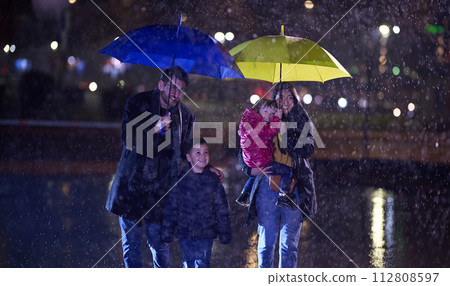 In the midst of a rainy urban night, a happy couple takes their children on a stroll through the city streets, heading towards the cinema for a delightful family movie outing 112808597