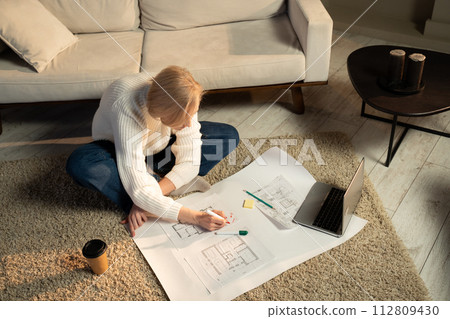 Young Woman Reviewing Floor Plans Beside Her Laptop in a Cozy Living Room 112809430