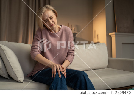Woman Experiencing Knee Pain While Sitting on Couch at Home Woman Experiencing Knee Pain While Sitting on Couch at Home 112809470