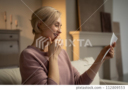 A woman smiles while reading a letter on her couch in the living room A woman smiles while reading a letter on her couch in the living room 112809553