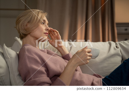 Mature Woman Relaxing on a Sofa With a Cup of Tea in a Cozy Room Mature Woman Relaxing on a Sofa With a Cup of Tea in a Cozy Room 112809634