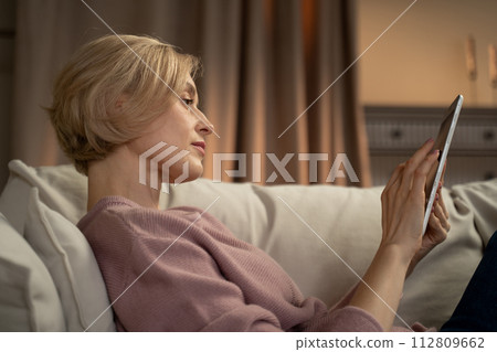 Mature Woman Enjoying a Tablet Session in Her Cozy Evening Living Room Mature Woman Enjoying a Tablet Session in Her Cozy Evening Living Room 112809662