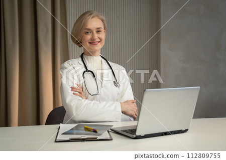 Confident Female Doctor in White Coat Sitting at Desk With Laptop in Office Setting Confident Female Doctor in White Coat Sitting at Desk With Laptop in Office Setting 112809755