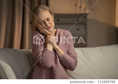 Mature Woman Sitting on Couch With Expression of Discomfort Due to Toothache 112809788