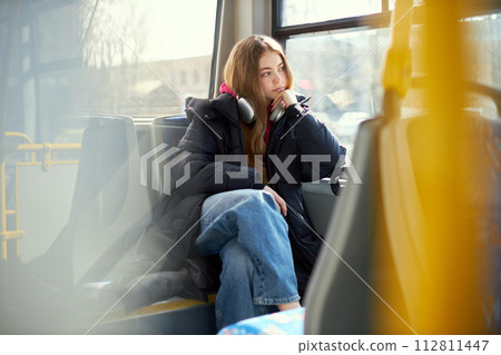 Dreamy looking young woman in headphones sitting in modern tram. Safe transportation. Blurred city streets on background 112811447