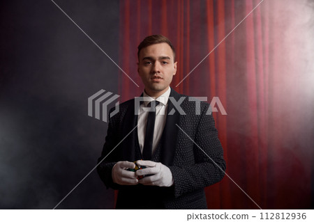 Magician wearing suit standing with cane on stage decorated red velvet curtain 112812936