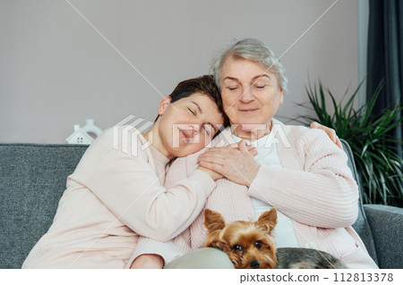 Smiling neutral gender woman and senior mother hugging, elderly mother and adult daughter enjoying tender moment, hugging, holding hands, happy family spending leisure time at home together 112813378