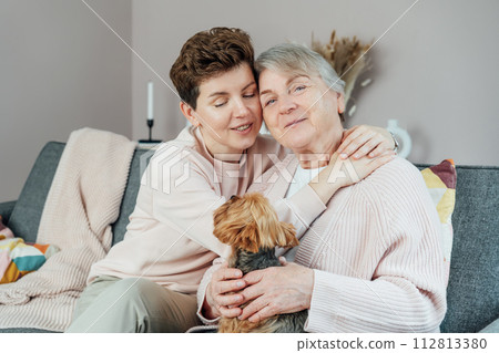 Portrait of happy senior 70s mother, adult neutral gender daughter and Yorkshire terriers dog hugging. Happy family enjoying weekend together in living room. Spending quality time in retiring house 112813380