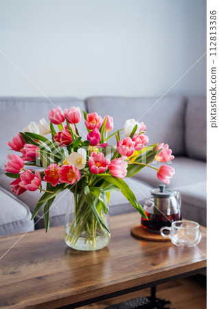 Focus on vase of fresh tulips bouquet and just brewed tea pot and cup on coffee table with gray couch sofa in modern leaving room. Cozy, comfortable relax at home. Time to take a break. Vertical card. 112813386