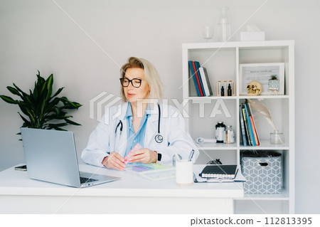 Friendly female middle aged doctor shows pills organizer to patient on laptop in clinic office. Video call online, treatment remote, pharmacy, supplement advice, medical healthcare. Telemedicine 112813395