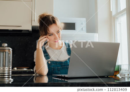Remote work at home. Concentrated young woman working on Laptop at the Kitchen Table in tiny flat apartments. A female student having online classes, Freelancer working from home. Online Job. 112813409