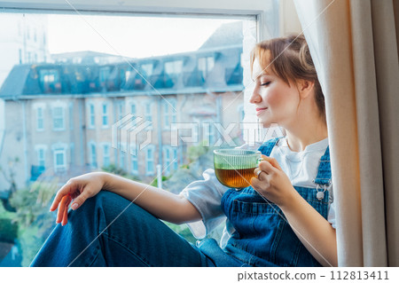 Time To Relax. Young woman looks out the window overlooking the city, sits on the windowsill at cozy home holds cup of hot tea drink. Happy calm female taking break for mental health wellbeing. 112813411
