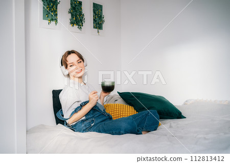 Smiling Woman listen music, dancing and enjoy cup of coffee or tea. Calm female spend free time at home enjoy favorite song with wireless modern headphones. Pastime weekend relax, no stress concept Smiling Woman listen music, dancing and enjoy cup of coffee or tea. Calm female spend free time at home enjoy favorite song with wireless modern headphones. Pastime weekend relax, no stress concept 112813412