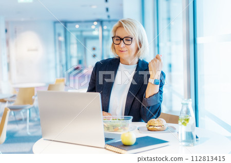 Middle-aged business woman working on laptop and having healthy lunch at working place, office cafe. Alternative office lifestyle female working in video call conference during lunch time. Modern job 112813415