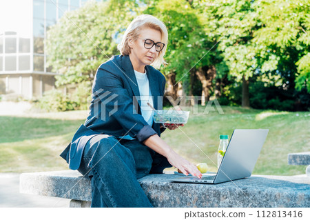 Business woman eating fast and healthy in front of laptop during job lunch break outdoors. Modern business lifestyle. Middle aged lady eats salad and use notebook pc on bench. Balanced diet lunch box. Business woman eating fast and healthy in front of laptop during job lunch break outdoors. Modern business lifestyle. Middle aged lady eats salad and use notebook pc on bench. Balanced diet lunch box. 112813416