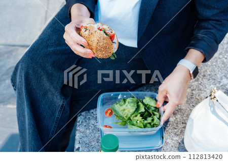 Top view close up business woman eating sandwich and fresh salad on lunch break sitting on bench at park. Balanced diet lunchbox for weight loss. Healthy eating habits and well-being. Selective focus. Top view close up business woman eating sandwich and fresh salad on lunch break sitting on bench at park. Balanced diet lunchbox for weight loss. Healthy eating habits and well-being. Selective focus. 112813420