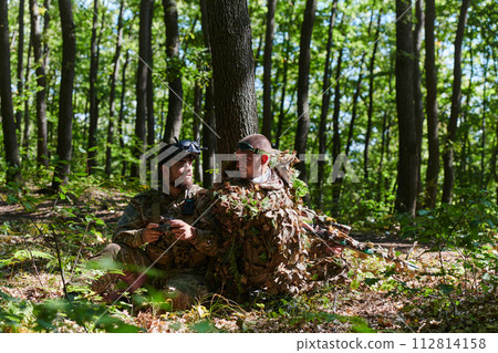 A skilled sniper and a soldier operating a drone with VR goggles strategize and observe the military action while concealed in the forest 112814158