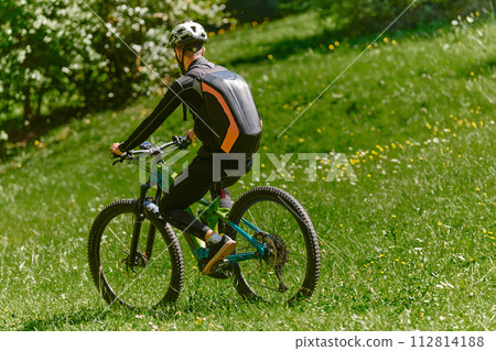 In the radiant glow of a sunny day, a fitness enthusiast, donned in professional gear, pedals through the park on his bicycle, embodying strength and vitality in a dynamic outdoor workout 112814188
