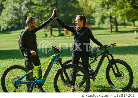 A sweet couple, adorned in cycling gear, rides their bicycles, their hands interlocked in a romantic embrace, capturing the essence of love, adventure, and joy on a sunlit path 112814201