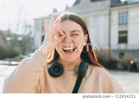 Happy young 30s woman showing ok sign with fingers over face on urban background, spring time. Human emotions, facial expression concept Happy young 30s woman showing ok sign with fingers over face on urban background, spring time. Human emotions, facial expression concept 112814231