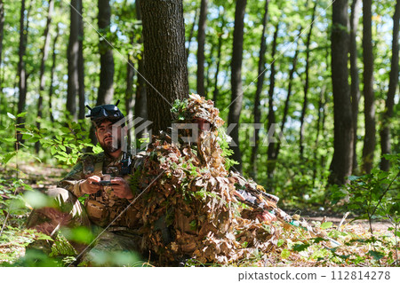 A skilled sniper and a soldier operating a drone with VR goggles strategize and observe the military action while concealed in the forest 112814278
