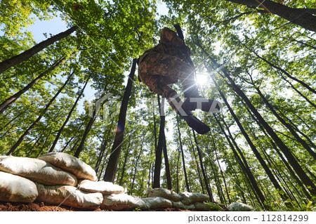 An elite soldier adeptly clears military barriers in the perilous wooded terrain, showcasing tactical skill and agility during specialized training 112814299