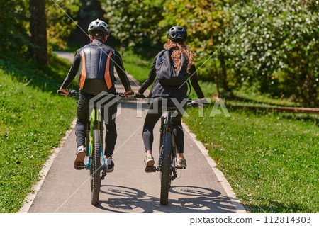 A blissful couple, adorned in professional cycling gear, enjoys a romantic bicycle ride through a park, surrounded by modern natural attractions, radiating love and happiness A blissful couple, adorned in professional cycling gear, enjoys a romantic bicycle ride through a park, surrounded by modern natural attractions, radiating love and happiness 112814303