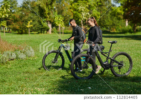 A blissful couple, adorned in professional cycling gear, enjoys a romantic bicycle ride through a park, surrounded by modern natural attractions, radiating love and happiness A blissful couple, adorned in professional cycling gear, enjoys a romantic bicycle ride through a park, surrounded by modern natural attractions, radiating love and happiness 112814405