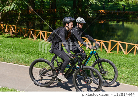 A blissful couple, adorned in professional cycling gear, enjoys a romantic bicycle ride through a park, surrounded by modern natural attractions, radiating love and happiness 112814444