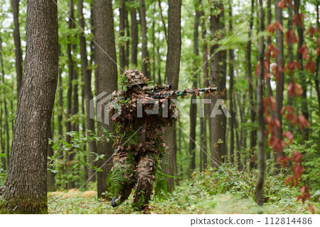 A highly skilled elite sniper, camouflaged in the dense forest, stealthily maneuvers through dangerous woodland terrain on a covert and precise mission 112814486