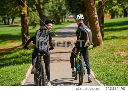 A blissful couple, adorned in professional cycling gear, enjoys a romantic bicycle ride through a park, surrounded by modern natural attractions, radiating love and happiness A blissful couple, adorned in professional cycling gear, enjoys a romantic bicycle ride through a park, surrounded by modern natural attractions, radiating love and happiness 112814498