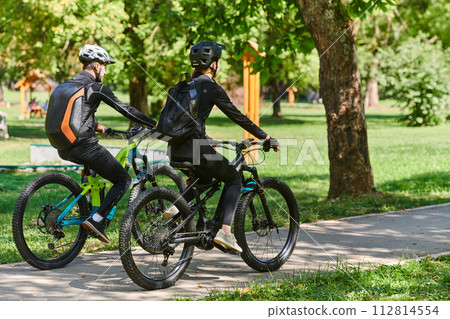 A blissful couple, adorned in professional cycling gear, enjoys a romantic bicycle ride through a park, surrounded by modern natural attractions, radiating love and happiness 112814554